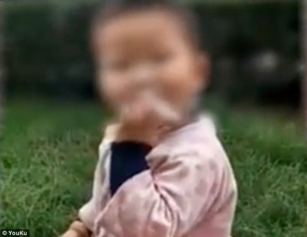 A toddler boy is seen taking puffs off a lit cigarette as a man and a woman film him in 
