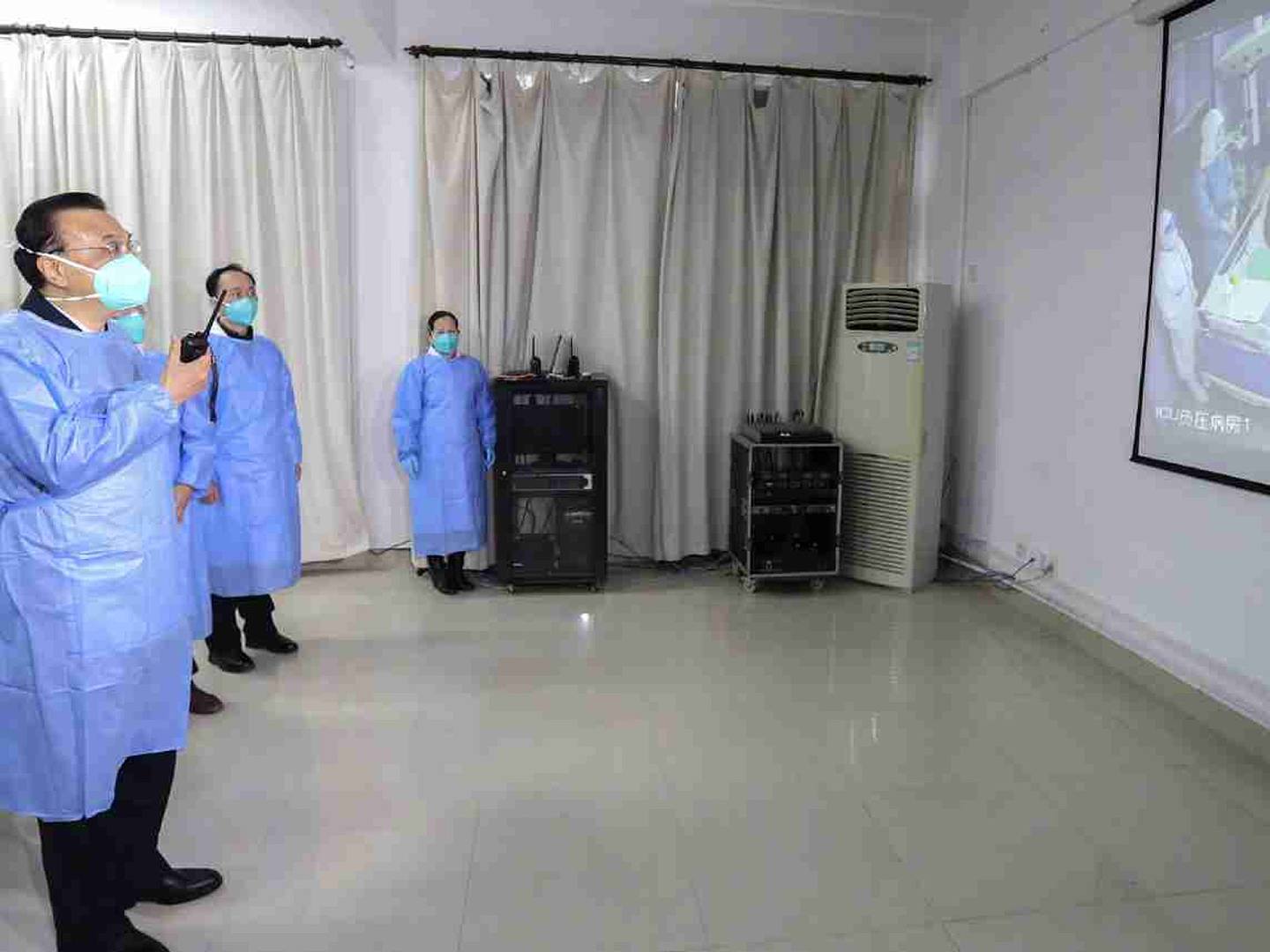 1月27日，李克强（左）在武汉市一家医院身穿防护服、戴口罩，通过投影仪观察医护人员救治病人情况。（中国政府网）