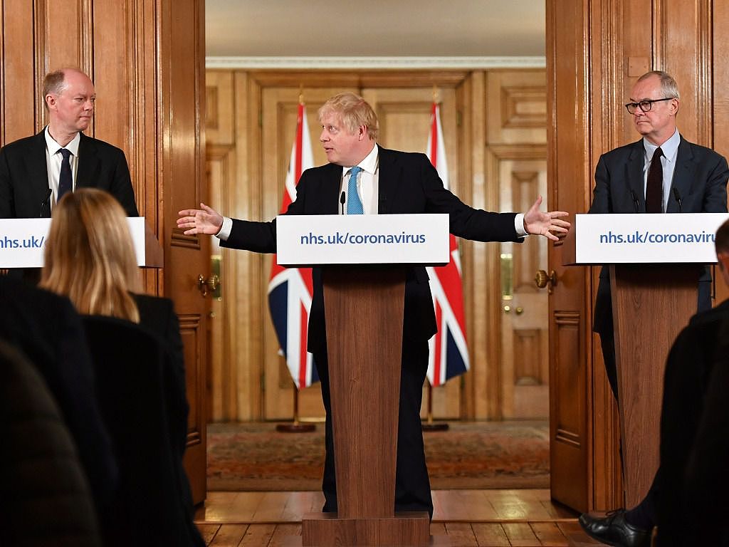 英国首相约翰逊（中）在3月19日于唐宁街10号举行的一次冠状病毒新闻发布会上，向首席医疗官惠蒂教授（左）和首席科学顾问瓦兰斯（右）手势。正是瓦兰斯提出了“集体免疫”的想法。（美联社）