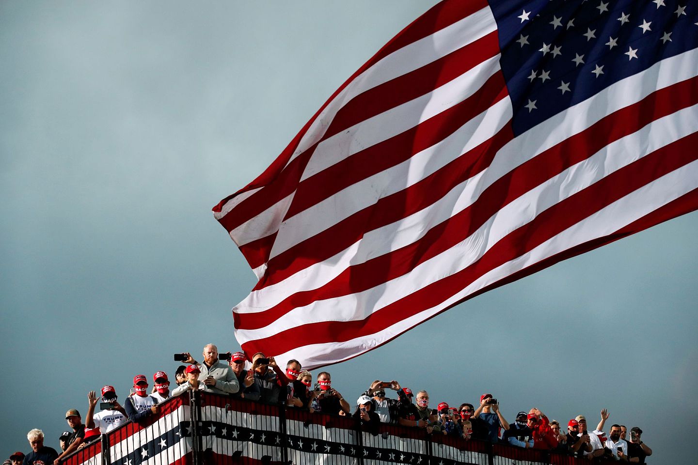 2020年10月14日，特朗普在艾奥瓦州得梅因国际机场（Des Moines International Airport）举行竞选活动，戴着“让美国再次伟大”帽子的支持者在一旁注视着美国国旗飘扬。（Reuters）