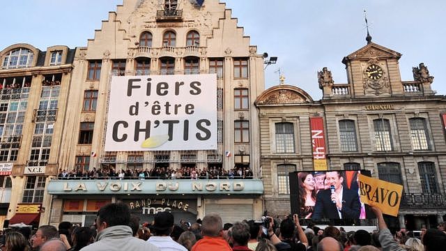 Town hall in northern France with crowd and large banner reading 'Proud to be from the Sticks'