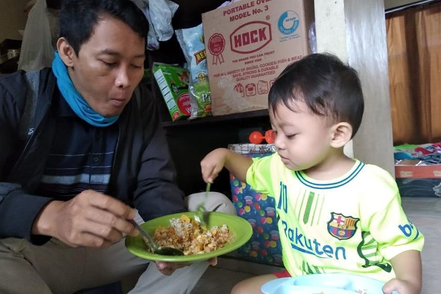 An Asian man sits on a small stool next to his young son, who has his spoon in his father's bowl of food. 