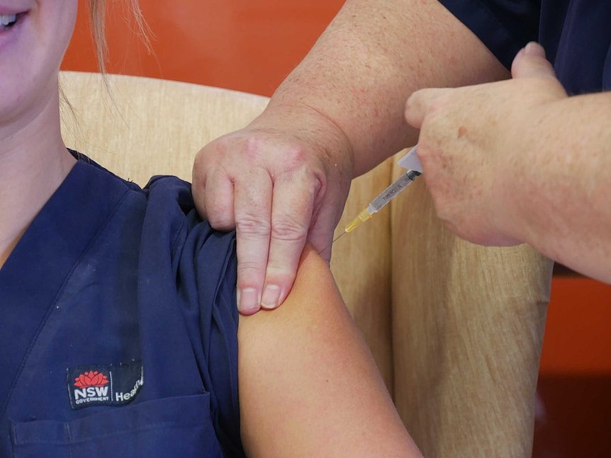 A syringe is inserted into the left shoulder of a uniformed female nurse, who is seated and smiling.