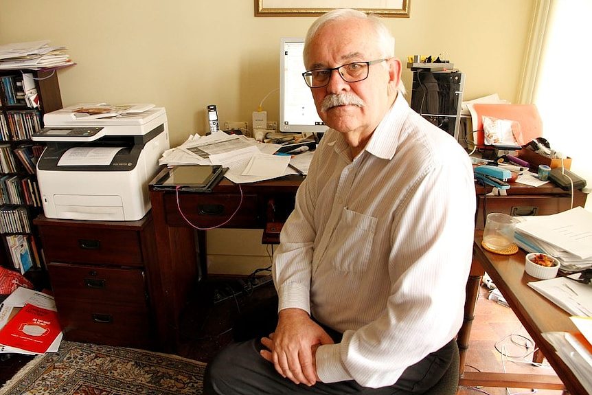 Dr Stephen Duckett sits at a desk cluttered with papers, books and stationary and a computer.