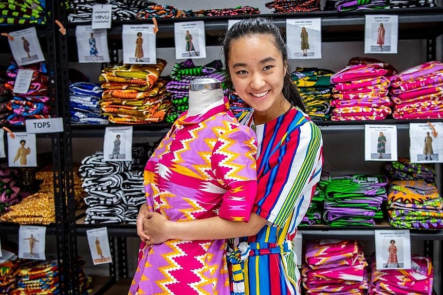 Stephanie Trinh-Tran hugs a mannequin dressed in brightly coloured clothes, while smiling at the camera.