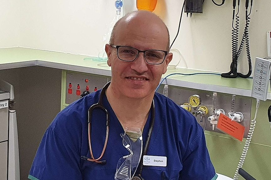 A man in a blue shirt and blue trousers sits on a hospital bed with a stethoscope around his neck and hospital equipment behind