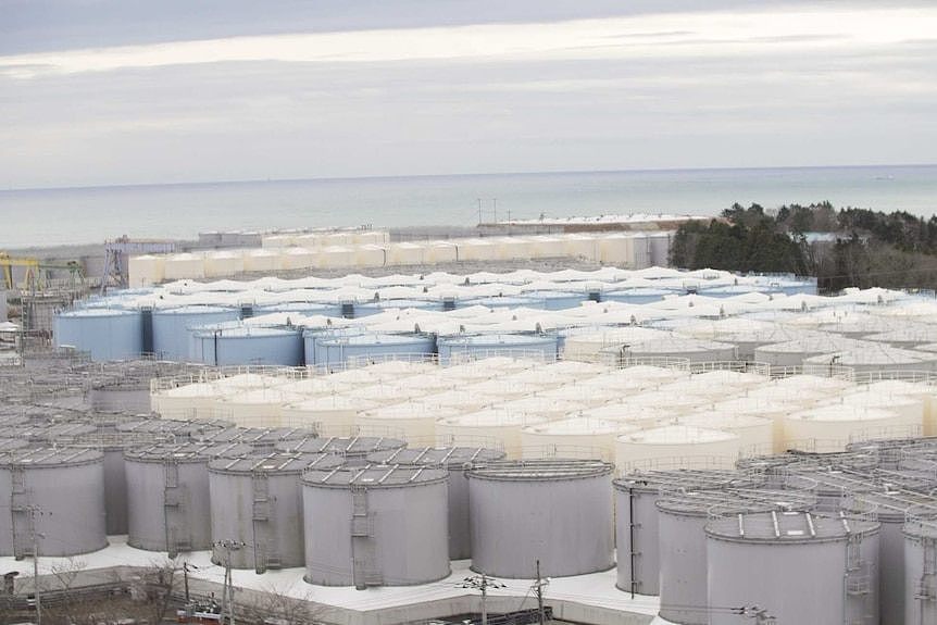 A group of grey and white tanks sit side by side near a body of water.