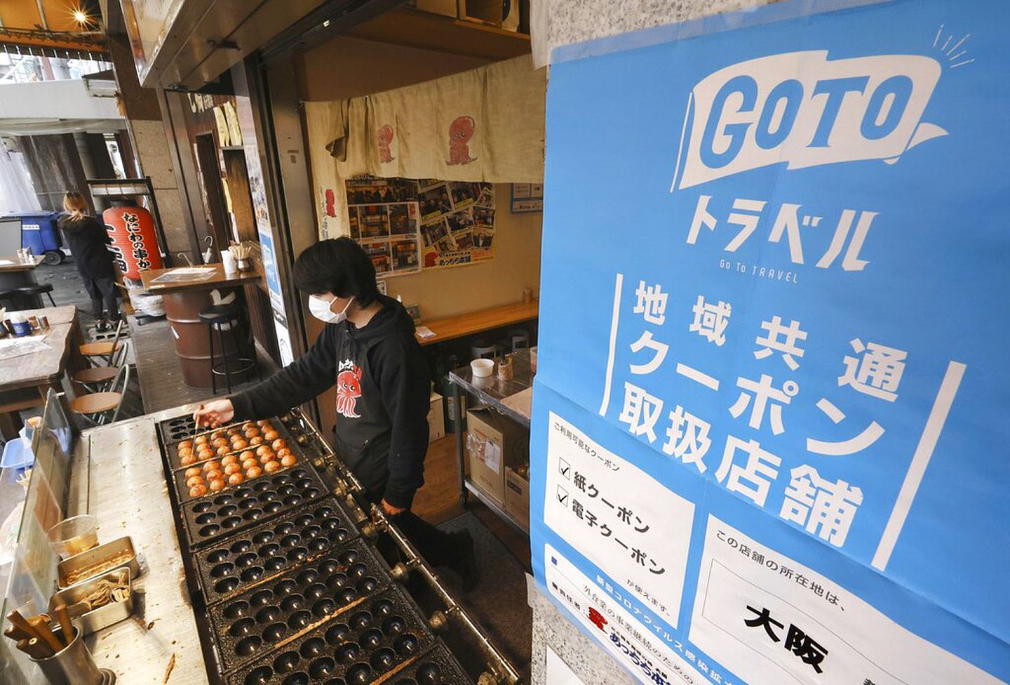 2021年12月28日大阪一个发售烤丸子的小贩，他的档摊张贴菅义伟推广的日本振兴旅游资助计划“去旅游吧”（GoTo Travel）的海报，计划在12月28日暂停。（AP）