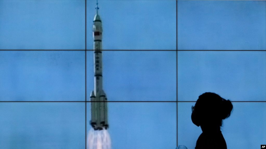 在北京，一名戴口罩的女子从电视屏幕前走过，电视屏幕上播放着搭载神舟十二号载人飞船的长征二号F遥十二运载火箭从酒泉卫星发射中心升空的景象。（2021年6月17日）