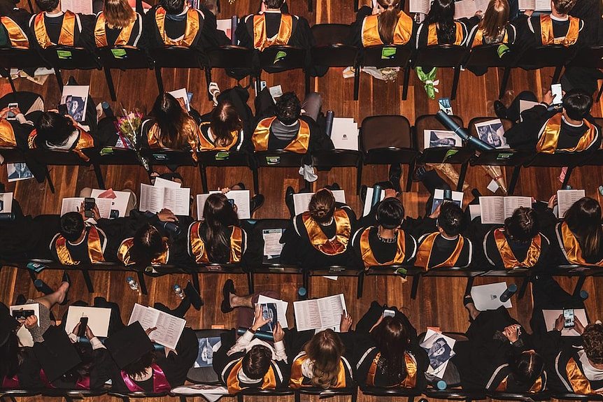 An aerial view of rows of seated graduation students with colourful sashes over their black academic robes.