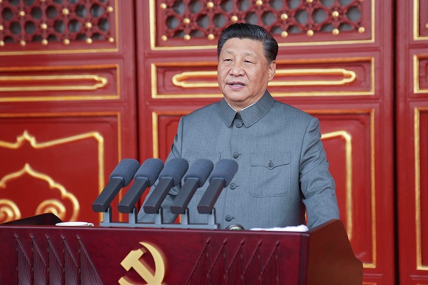 Xi Jinping delivers a speech in front of red doors and a podium with a sickle and hammer.