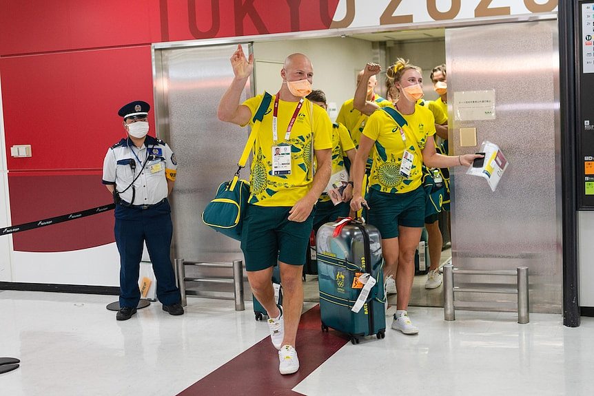 Australian athletes in green and gold tracksuits and face masks wave at an airport terminal 