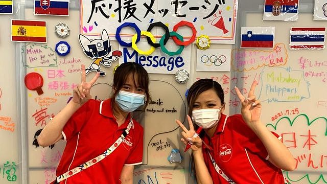 Two volunteers pose behind a wall with handwritten messages to cheer on Olympic athletes