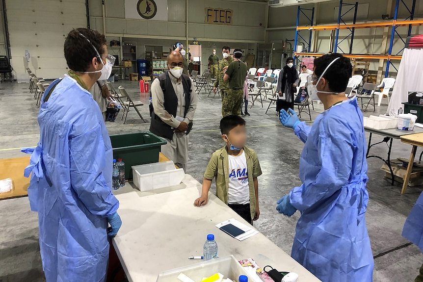 People wearing masks walk toward medical professionals wearing blue gowns and masks in a large hangar space