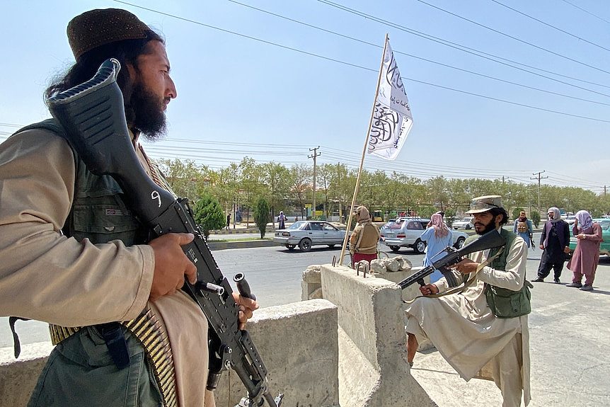 A Taliban fighter stands on top of a guard tower, holding a gun. A white flag is visible behind him