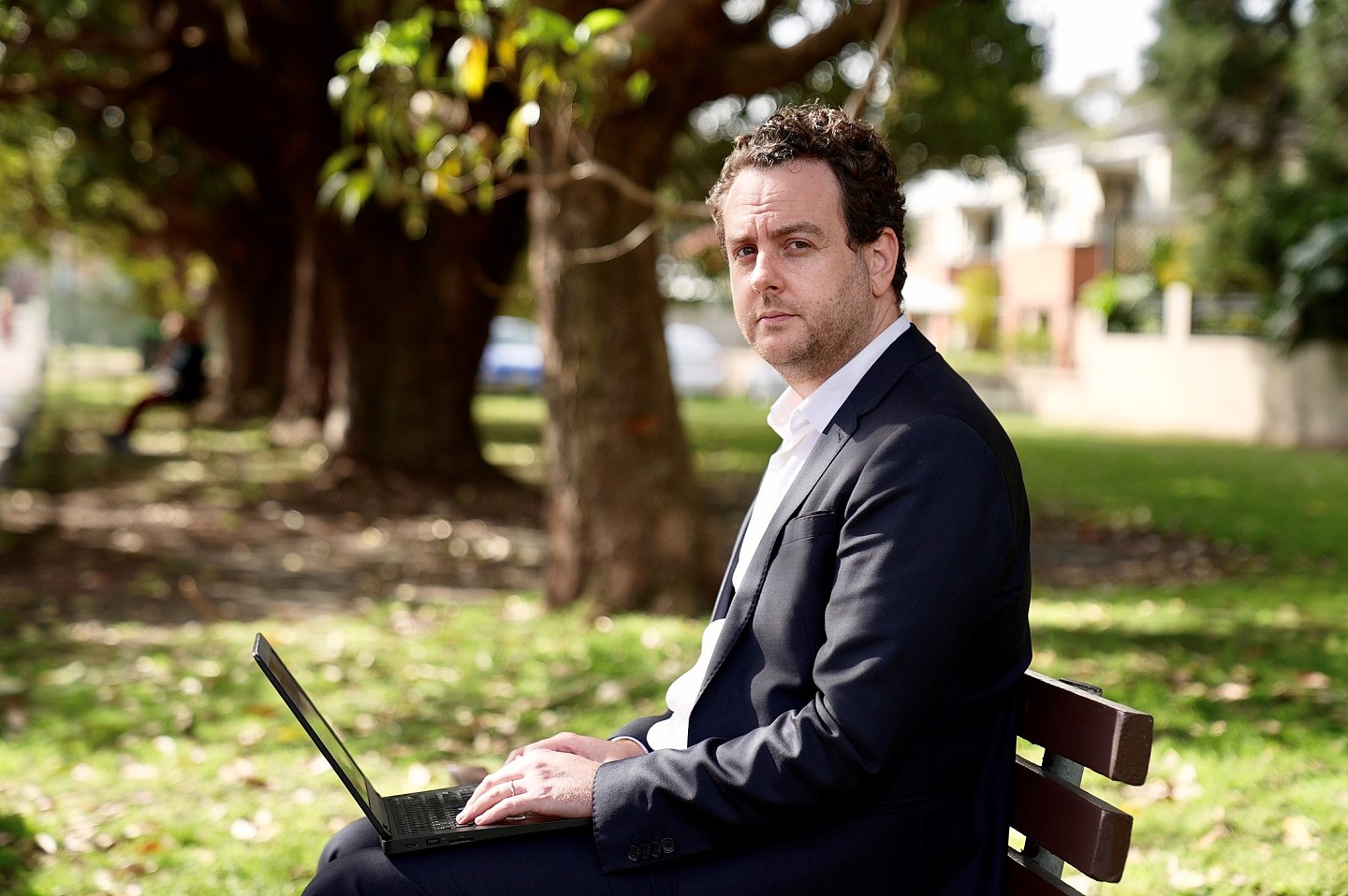 Xavier in a black suit sits at a park bench in Marrickville.