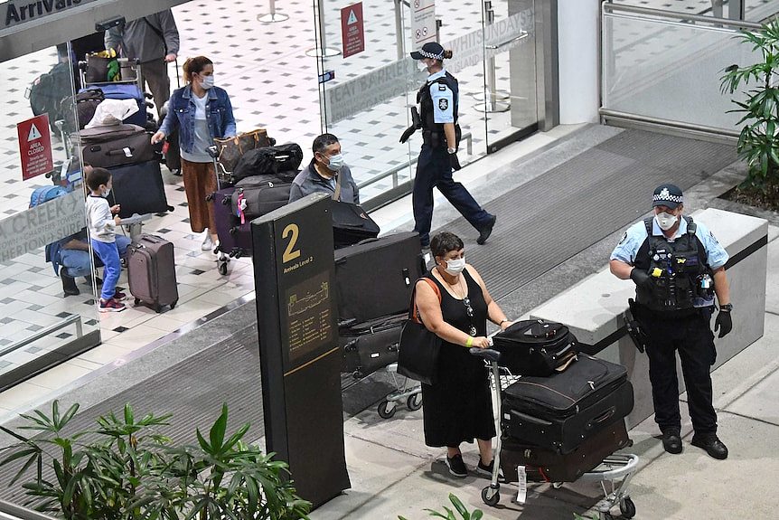 在警方的引导下，一排人推着行李车，戴着医用口罩走过机场