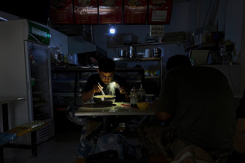 A man eats a bowl of noodles in a dark room, with only his phone light to illuminate what's in the bowl