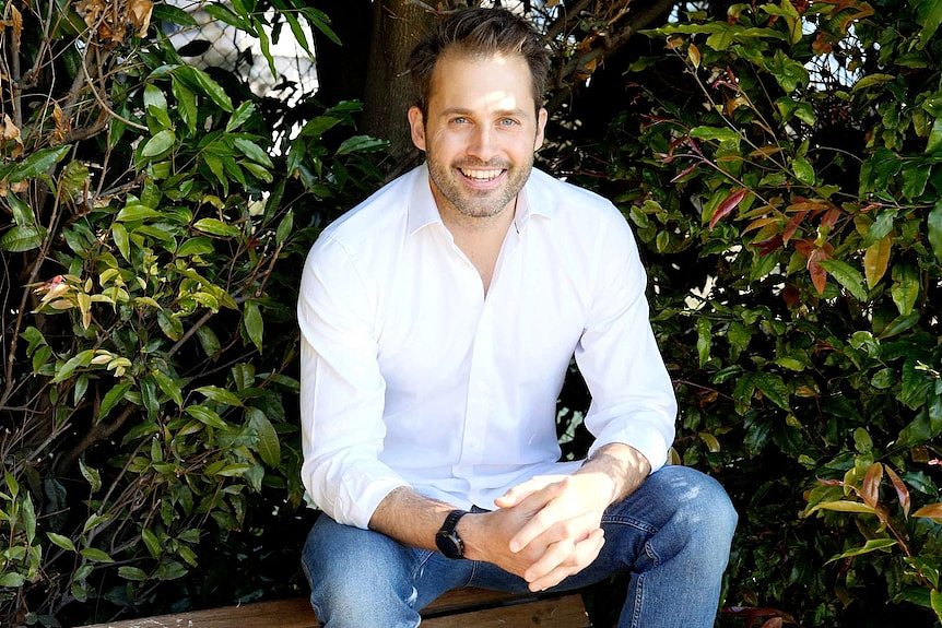 A smiling man in a white button up shirt and jeans sits atop a park bench as he poses for a photograph in front of green plants.