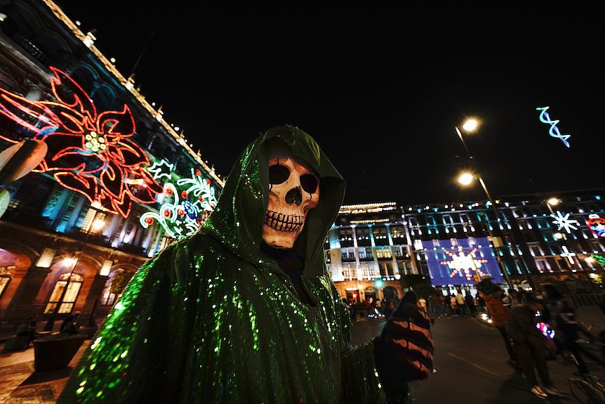 a person dressed in a skull mask and robe as folk saint la santa muerte poses in front of christmas lights