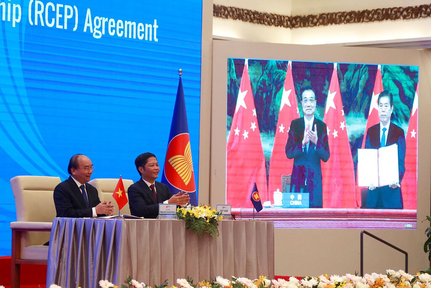2020年11月15日，越南总理阮春福（左一）与该国贸易部长陈俊英（左二）以视频形式在越南河内的东盟峰会。屏幕中鼓掌者是中国国务院总理李克强，身旁的是时任中国商务部长钟山。（AP）