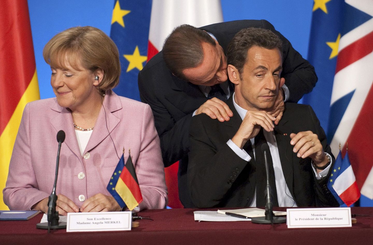 2008年10月4日，意大利总理贝卢斯科尼在巴黎参加欧洲应对全球金融危机峰会期间，与时任法国总统萨科齐（右）耳语，德国总理默克尔（左）发出笑声。（VCG）