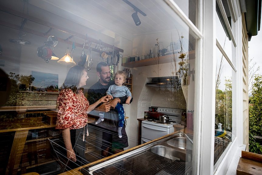 A mother and father play with their baby boy. The photo has been taken from outside through their kitchen window.