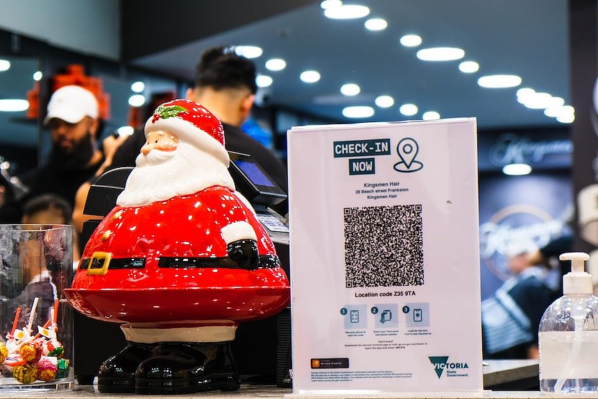 A tiny santa sits on a shop counter next to a QR check in board and a hand sanitizer and a jar of lollies.