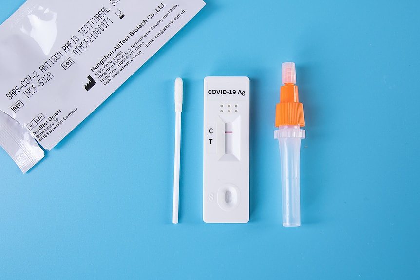A generic image of a Rapid Antigen Test packet, a swab, a small dropper bottle, and the test itself laid out on a blue surface.
