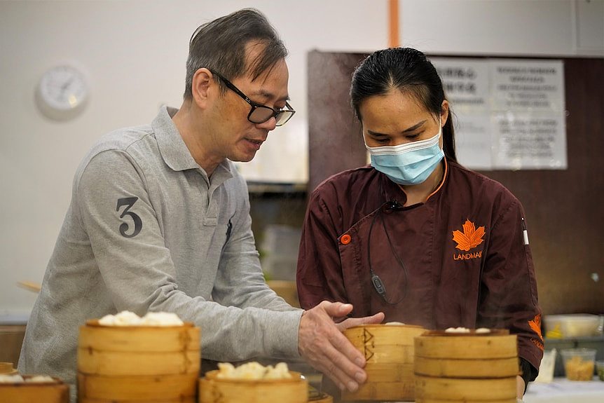 A man holds a bamboo box with dumplings as a woman with a masks watches.