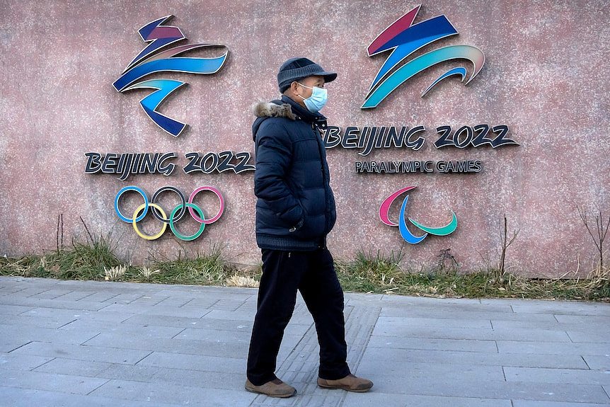 A man in a mask walks past two signs with Beijing 2022 logos featuring the Olympic rings