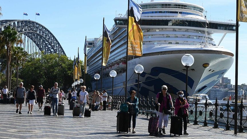 Several people walking past a large ship, as the Sydney Harbour Bridge can be seen in the background