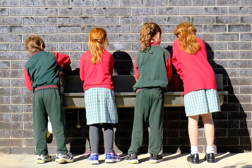 Four primary school children stand with their backs to the camera and wash their hands at a long metal basin on a brick wall.