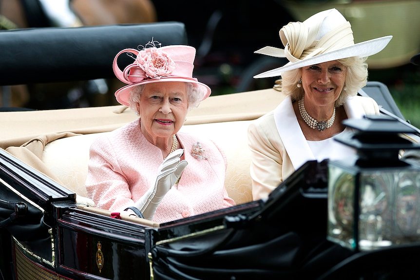 Queen Elizabeth and Camilla sit side by side in a carriage smiling and waving 