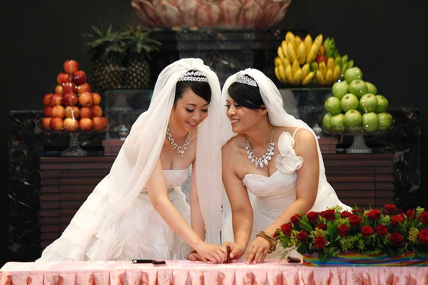 Two women wearing wedding dresses stamp their names in front of a statue of Buddha in a prayer hall.