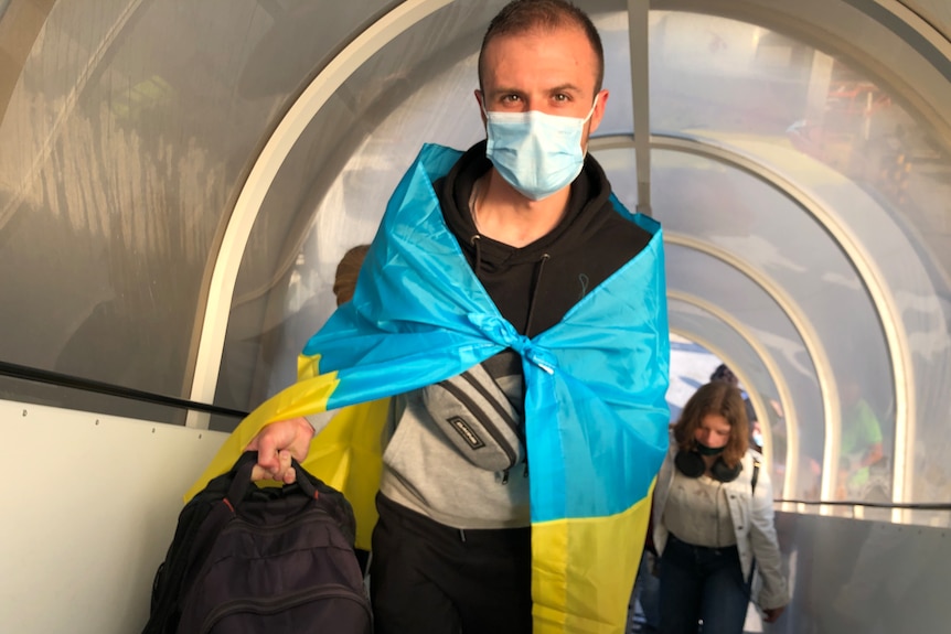 A man wearing a maks and draped in an Ukranian flag travels up an escalator