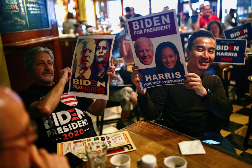 在台湾台北，一名男子手持印有美国民主党总统提名人乔-拜登和竞选伙伴卡玛拉-哈里斯参议员图像的海报，其他人在酒吧观看美国大选的现场结果。