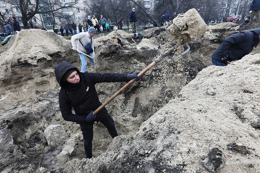 People use shovels to dig large holes in the ground, buildings and people in background. 