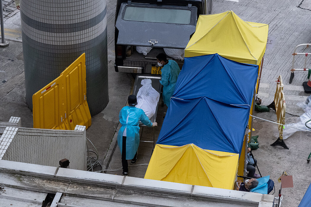 本月，香港殡仪馆的工作人员把一具尸体抬上车，旁边就是在一家医院外的临时治疗区接受治疗的人们。