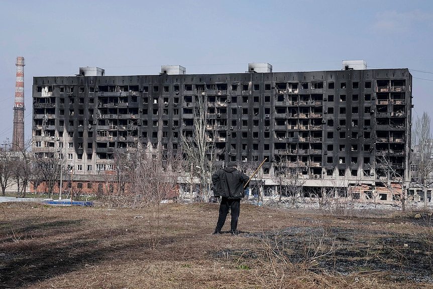 A man with a walking stick looks at a burned apartment building from a barren patch of land some distance away.