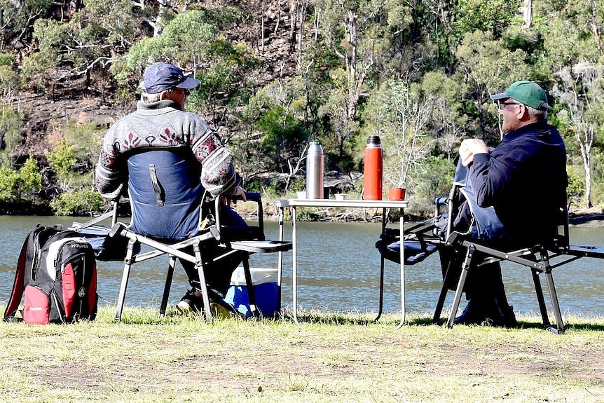 Two men in caps and jacket near a river talking as they sit on camping chairs and a table with a thermos.