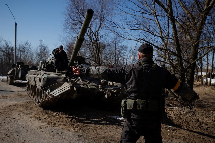 A soldier is seen from behind with his arms stretched out side to side, directing a tank