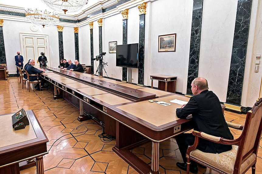 Putin, a man in a dark suit, sits at the head of a long table at which several people are sitting at the opposite end