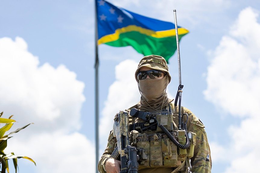 An ADF soldiers wearing a mask and sunglasses in uniform stands in front of a flag pole with the Solomon Islands flag