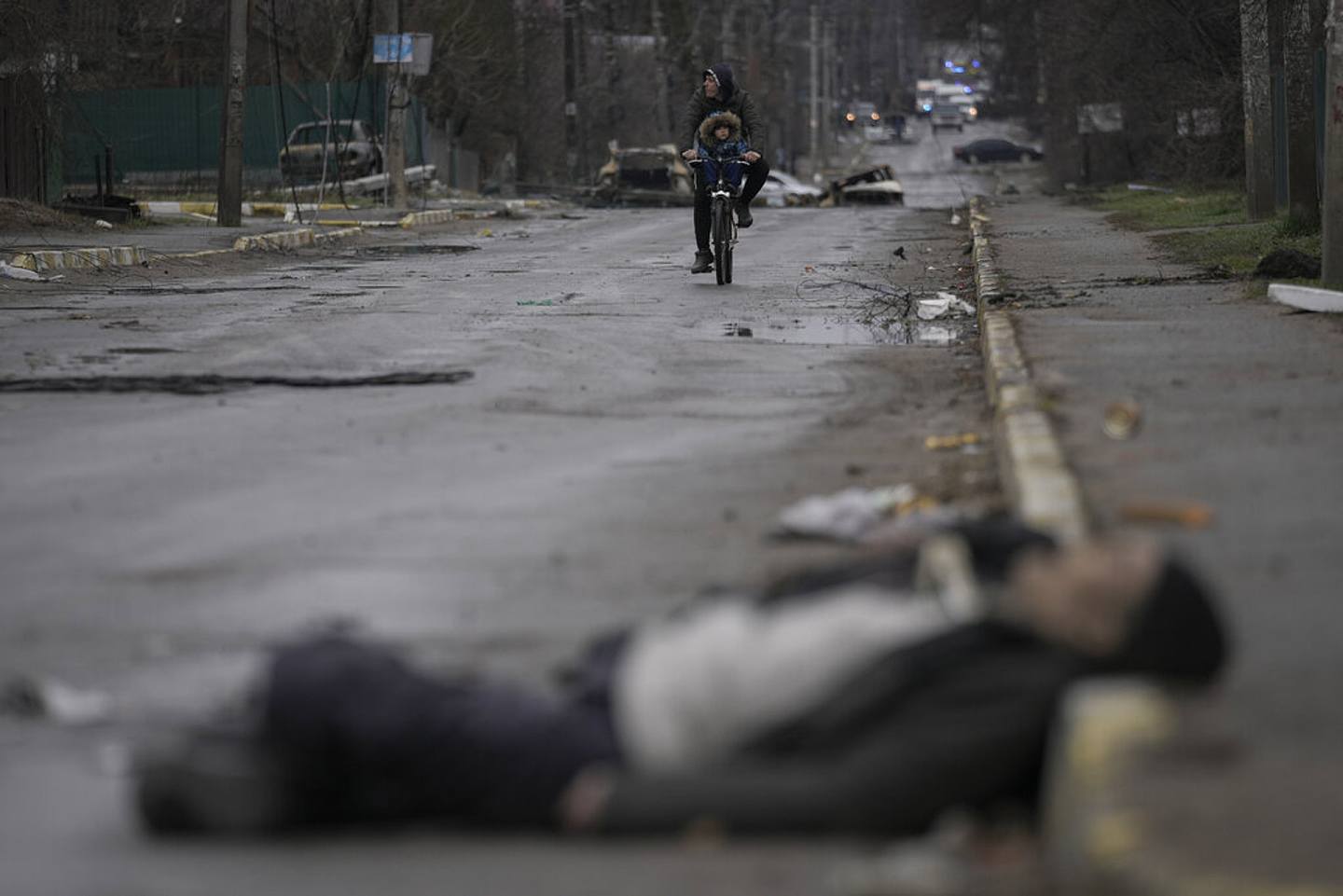 乌克兰局势：图为2022年4月2日一名男子与儿童骑单车经过布查镇的街头。 他们的前方有一具死者遗体。 （AP）