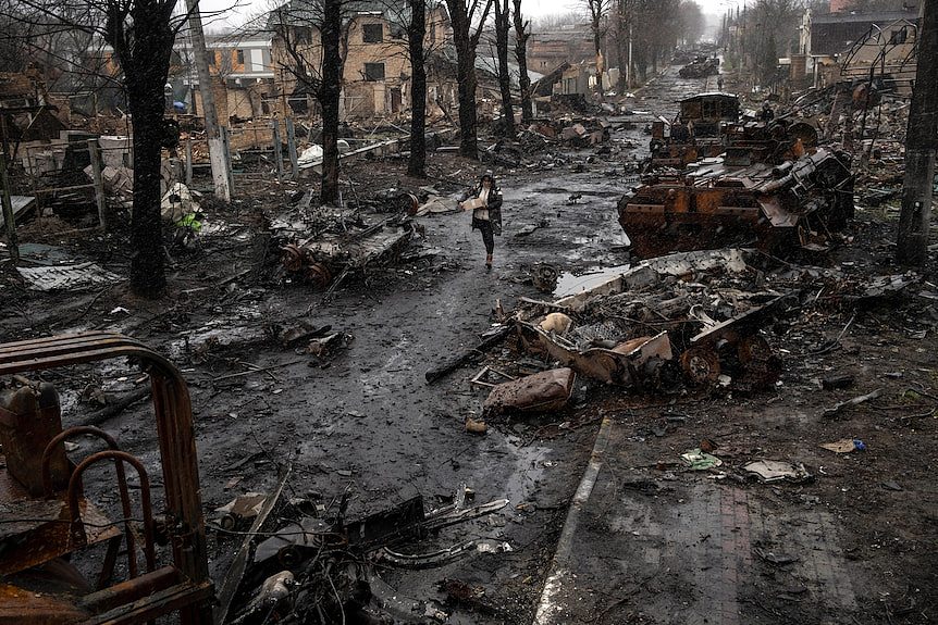 A woman carries goods as she moves between the wreckage of tanks on a residential street destroyed by war.