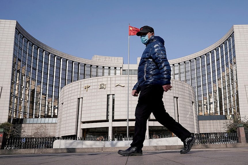 A man wearing a face mask and blue puffer jacket walks past a large building with a Chinese flag flying on a flagpole.