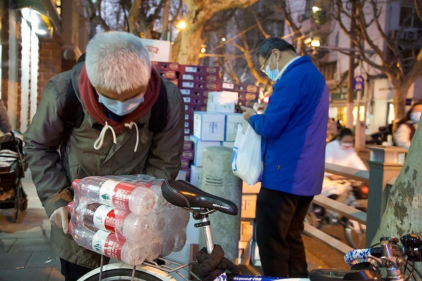 An old man is trying to put a bulk of bottled water in his bike basket. 