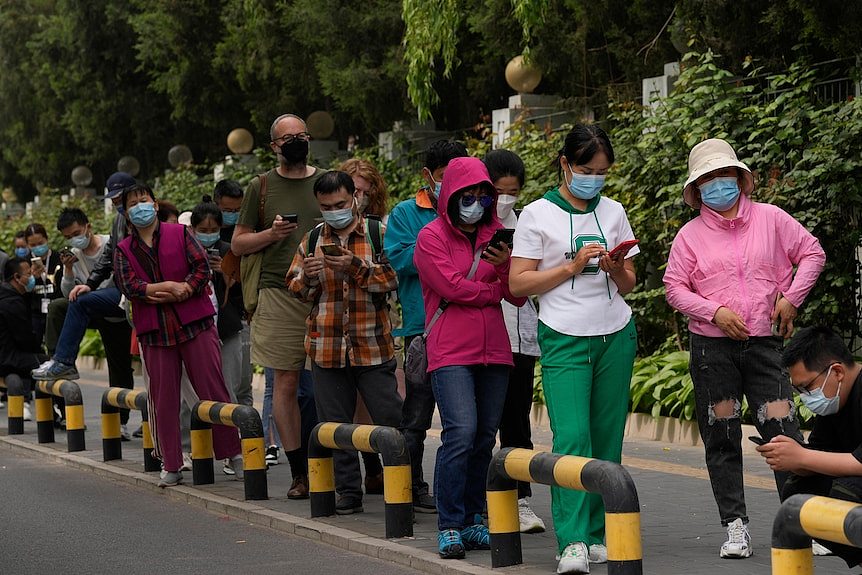 People in face masks stand in line on the street for a COVID-19 test. Many are looking at their phones.