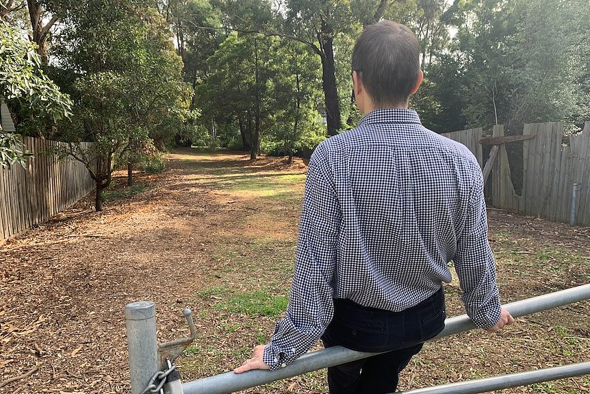 A man sitting on a metal barrier wearing a checked shirt looking towards trees.